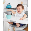 A happy baby in a high chair plays with a teal and grey feeding set, including stacking bowls, in a modern kitchen.