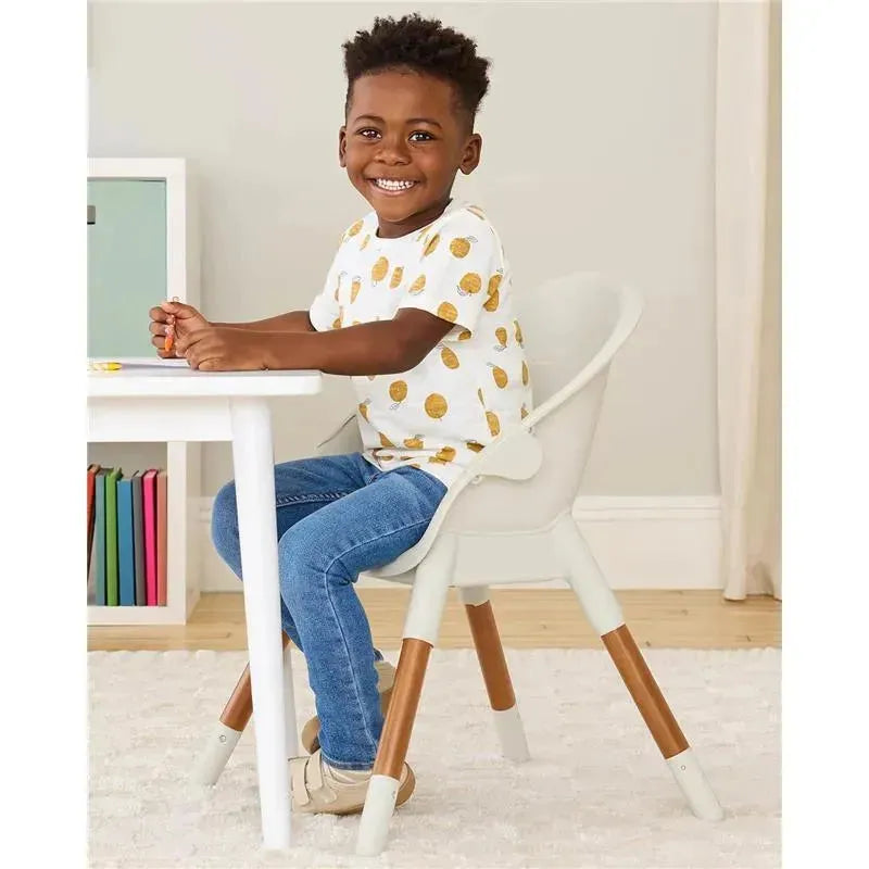 Child sitting in a Skip Hop EON 4-in-1 High Chair in Oat color, smiling while drawing at a white table.