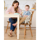 A woman and a smiling boy sit at a table featuring the Skip Hop EON 4-in-1 High Chair in oatmeal color, with food on the table.