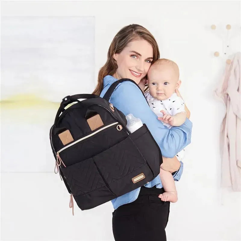 A woman holds a baby while showcasing the Skip Hop Suite Diaper Bag Backpack Set in black, featuring multiple compartments and a bottle holder.