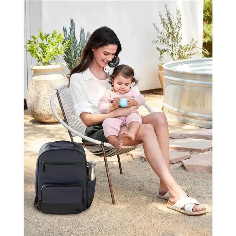 Navy Skip Hop Flex Sporty Diaper Bag Backpack beside a woman sitting with a toddler outdoors, holding a toy.