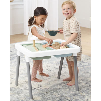 Two toddlers play at a Skip Hop Discoverosity 3-in-1 Toddler Sensory Table, filled with sand and playful tools.