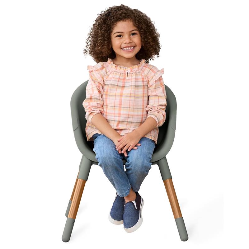 A smiling child with curly hair sits in the Skip Hop EON 4-in-1 Convertible High Chair, wearing a pastel plaid top and blue jeans.