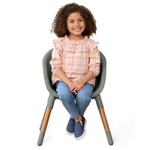 A smiling child with curly hair sits in the Skip Hop EON 4-in-1 Convertible High Chair, wearing a pastel plaid top and blue jeans.