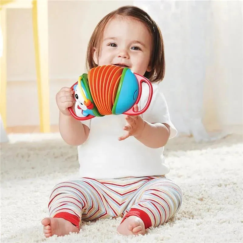 Toddler smiling while holding a colorful Skip Hop Hedgehog accordion toy, sitting on a soft rug.