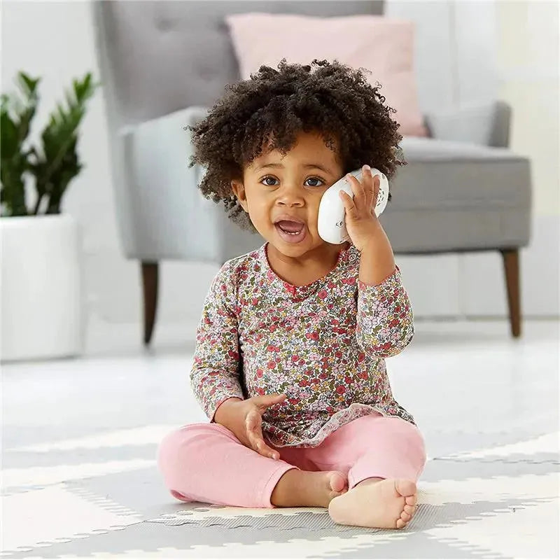 A toddler with curly hair sits on a mat, holding a white toy cellphone to her ear, smiling. She wears a floral long-sleeve top and pink pants.