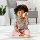 A toddler with curly hair sits on a mat, holding a white toy cellphone to her ear, smiling. She wears a floral long-sleeve top and pink pants.