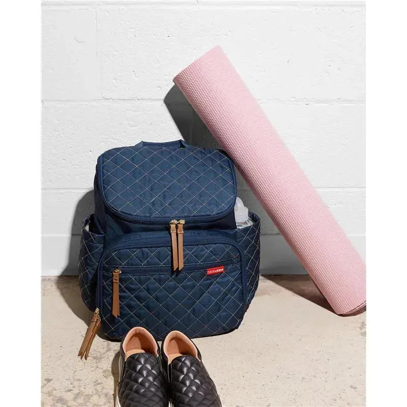 Navy Skip Hop Forma diaper bag backpack with quilted pattern, two zippers, and tan accents, alongside a pink yoga mat and black shoes.