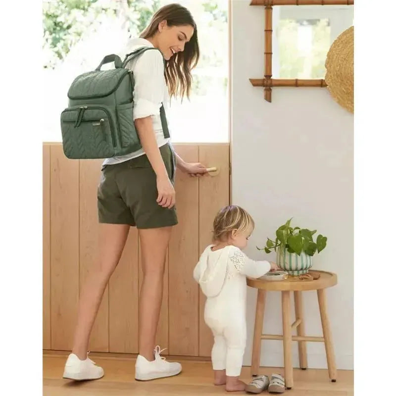 Sage Skip Hop Forma Diaper Backpack on a woman’s back as she opens a door, with a toddler at a nearby table exploring items.