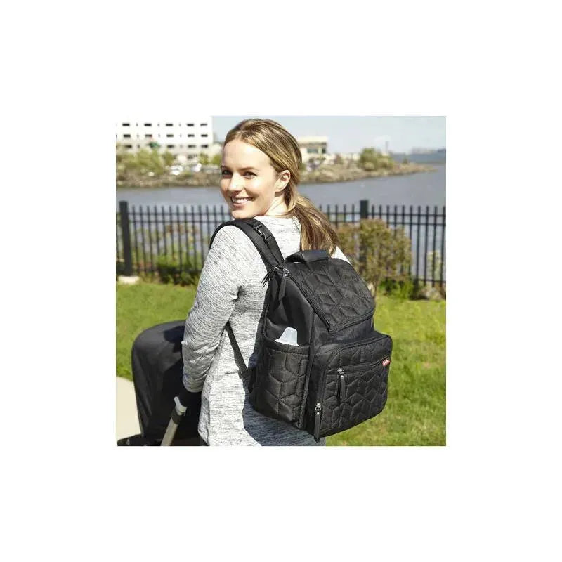 A woman smiles while wearing a black Skip Hop Forma Diaper Bag Backpack, showcasing its quilted design and side pockets.