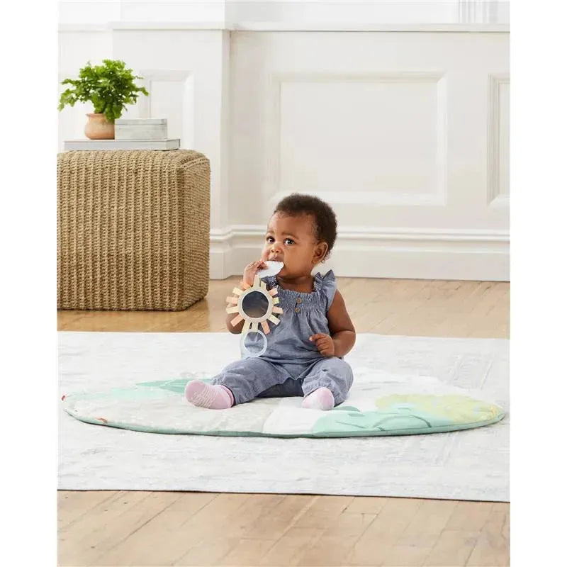 Baby sitting on a soft activity gym mat, holding a sun-shaped mirror toy. A plant and woven storage are in the background.