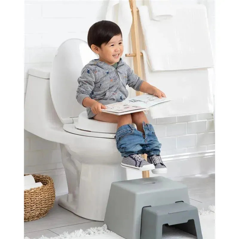 Child sitting on a white toilet reading a book, wearing a gray hoodie, with pants down and feet on a gray potty stool.