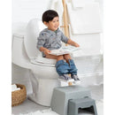 Child sitting on a white toilet reading a book, wearing a gray hoodie, with pants down and feet on a gray potty stool.