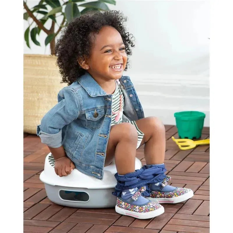 Child sitting on a Skip Hop Go Time 3-In-1 Potty, smiling, wearing a denim jacket and colorful shoes.