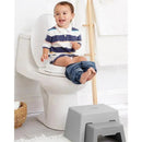 Toddler sitting on a toilet with pants around ankles, smiling, next to a gray potty stool for support.