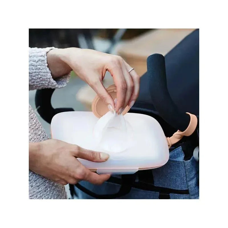 Hands reaching into a blush-colored Skip Hop Grab & Go Snug Seal Wipes Case, pulling out a moist wipe.