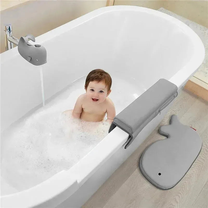 Baby sitting in a white bathtub filled with bubbles, smiling, with a grey whale-shaped faucet cover and a bath mat nearby.