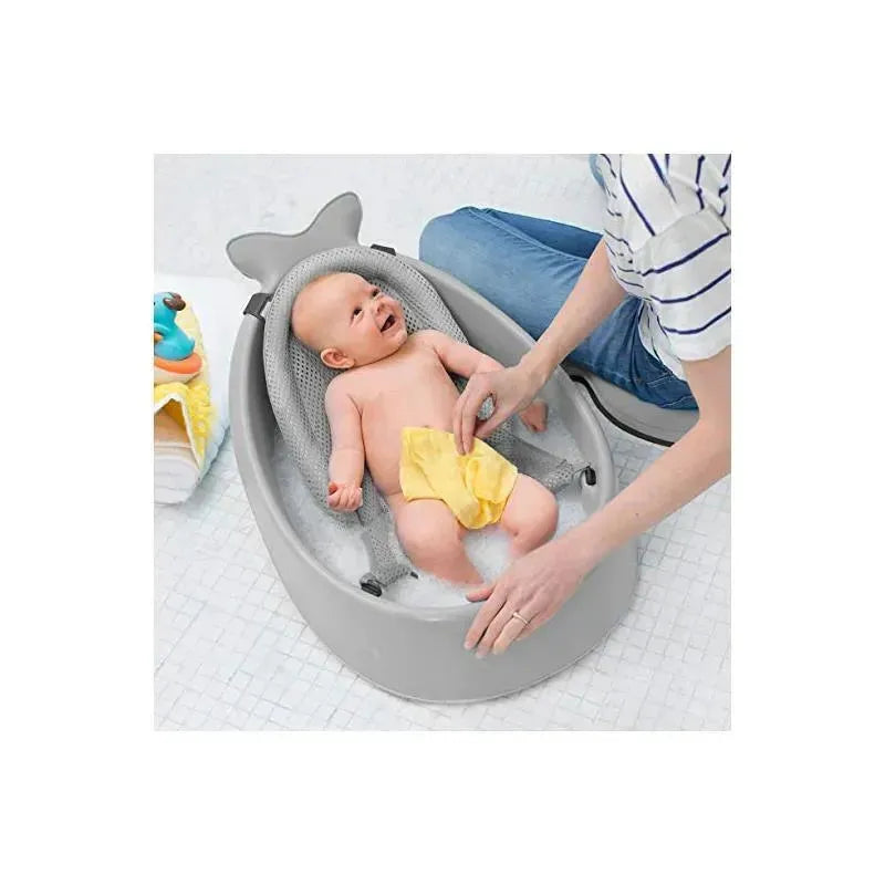 Skip Hop Moby Smart Sling 3-Stage Tub in grey with a baby wearing a yellow diaper being bathed, and a person assisting.