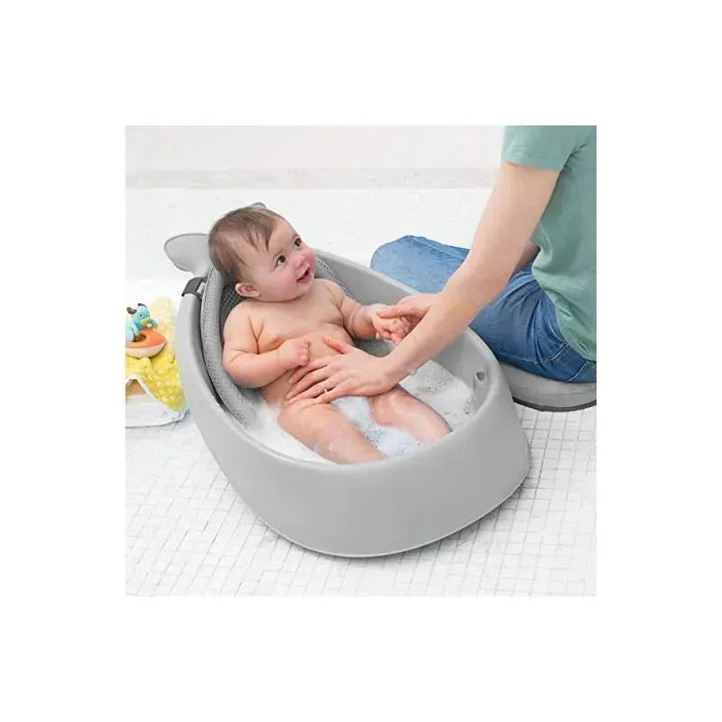 A baby is happily seated in a grey Skip Hop Moby Smart Sling 3-Stage Tub, with bubbles and a caregiver gently washing them.