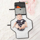 Portable baby changing pad in grey melange, featuring a child lying on it, with pockets for storage visible above.