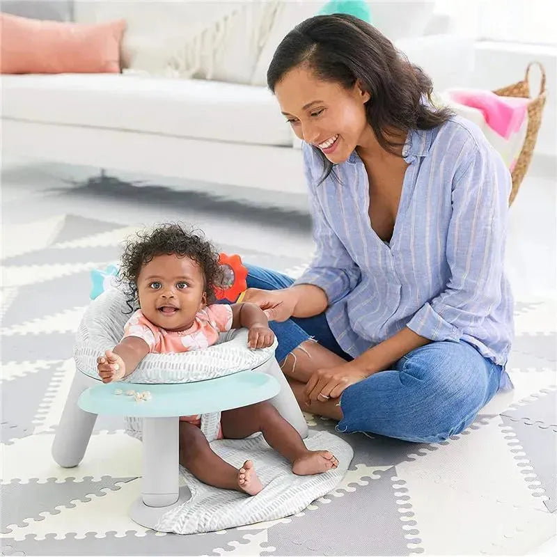 A baby with curly hair sits in a Skip Hop Silver Lining Cloud 2-in-1 Activity Floor Seat with a smiling woman beside them.