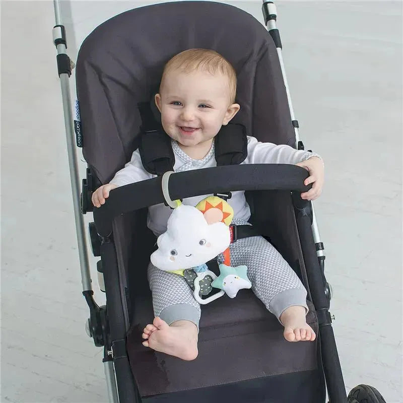 Baby smiling in a stroller, holding onto a Skip Hop Silver Lining Cloud Jitter Toy with colorful features and dangling characters.