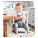 A happy toddler is seated on a grey and white Skip Hop Sleek Seat Booster, wearing a blue outfit with suspenders.