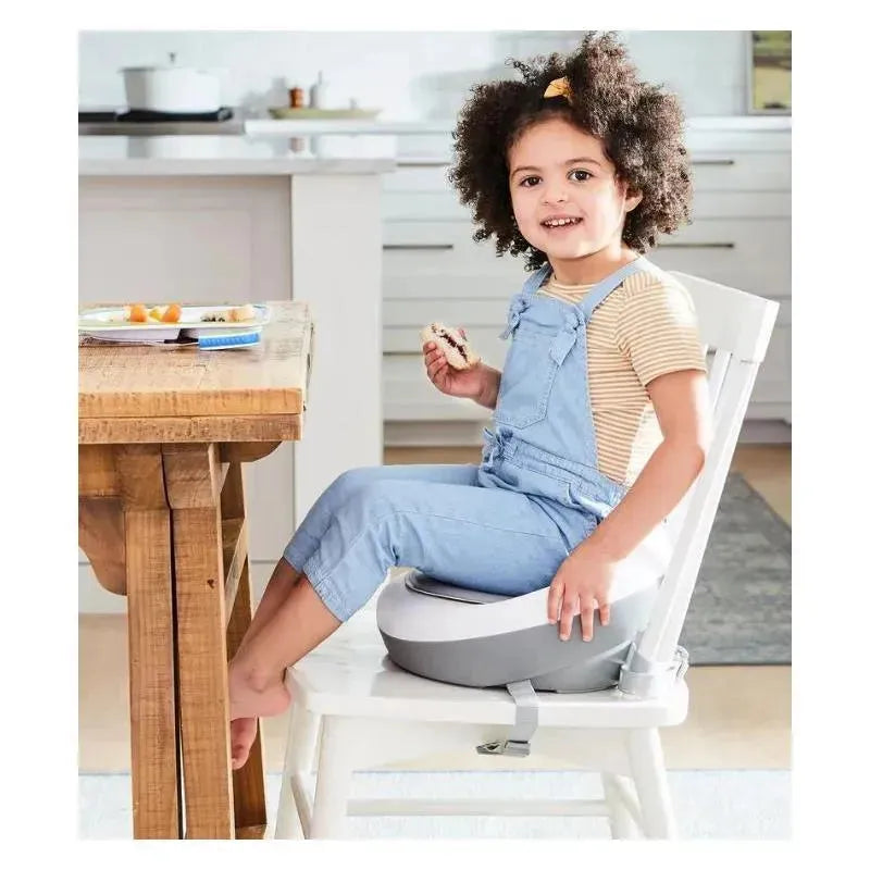 Child sitting on a grey and white Skip Hop Sleek Seat Booster at a kitchen table, holding a sandwich, smiling.