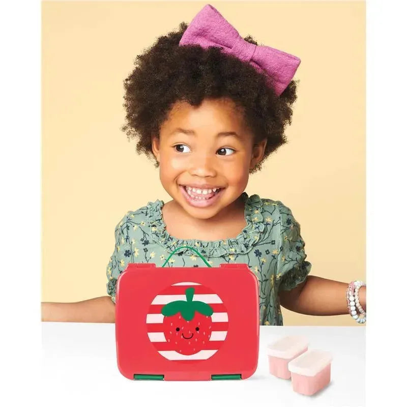Young girl smiling next to a red Skip Hop Bento Lunch Box featuring a strawberry design, with two pink food containers nearby.