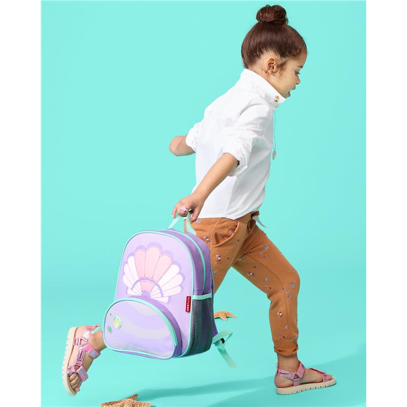 Little girl with a seashell-themed Skip Hop backpack, walking against a bright turquoise background.