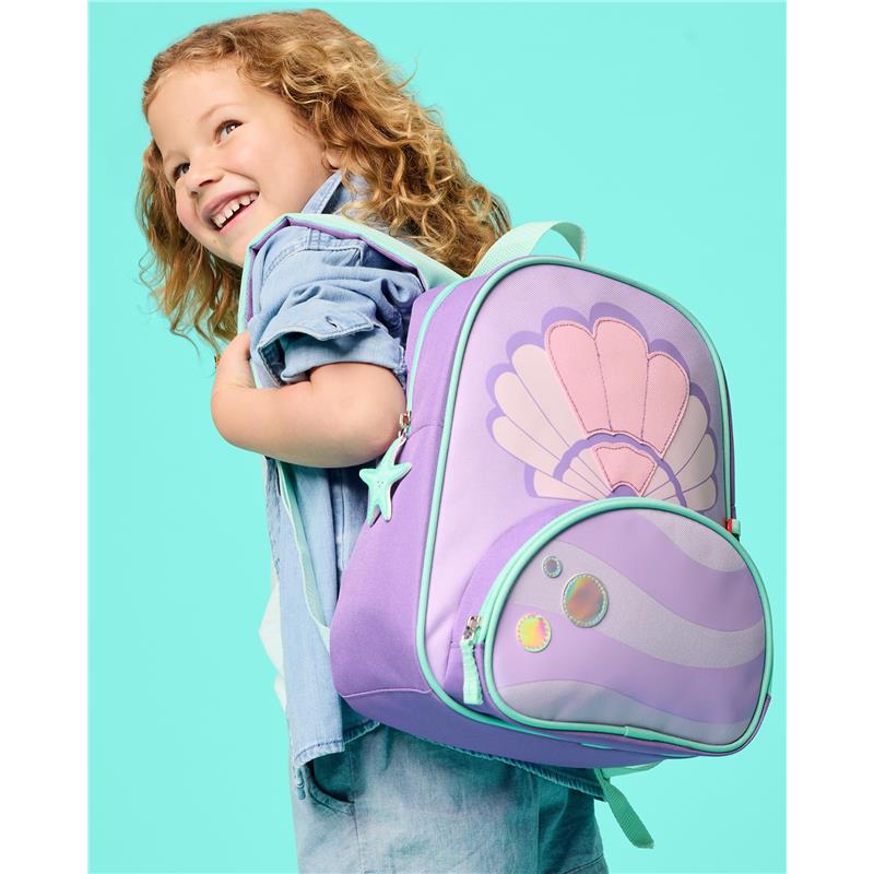 Little girl smiling while holding a seashell-themed purple backpack adorned with iridescent details.