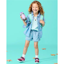 A smiling girl with curly hair wearing a denim shirt and shorts holds a Skip Hop stainless steel straw bottle decorated with seashells.