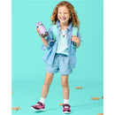 A smiling girl with curly hair wearing a denim shirt and shorts holds a Skip Hop stainless steel straw bottle decorated with seashells.