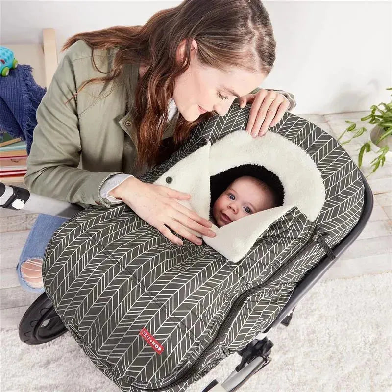 A woman gently holds a baby peeking out of a grey Skip Hop car seat cover with a feather pattern, featuring a soft white lining.