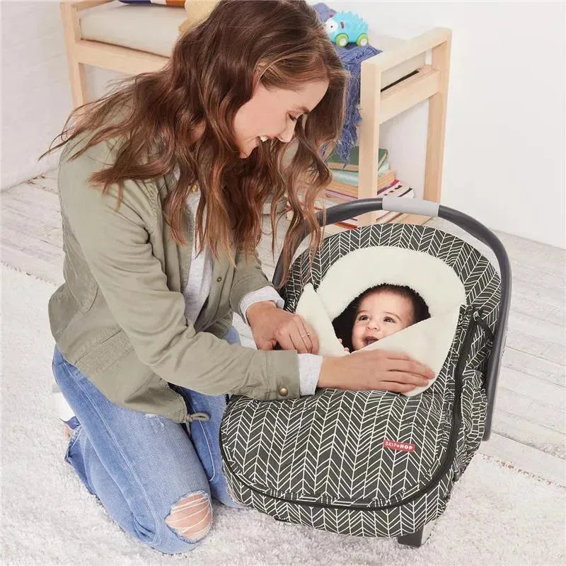 A mother is smiling while adjusting the Skip Hop Stroll & Go car seat cover, featuring a grey feather pattern, around her baby.