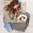A mother is smiling while adjusting the Skip Hop Stroll & Go car seat cover, featuring a grey feather pattern, around her baby.