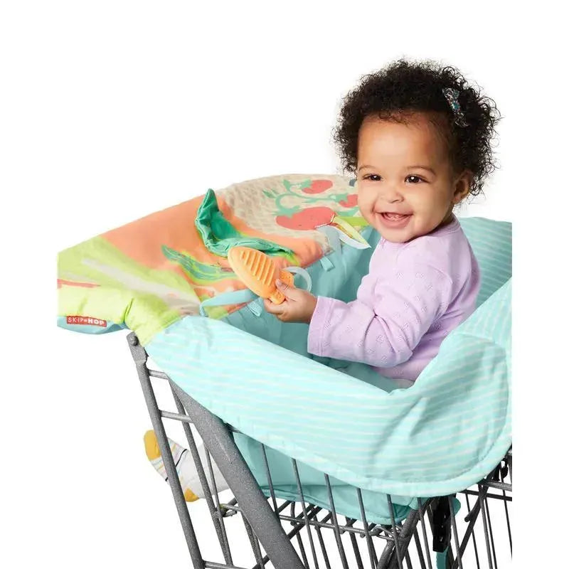 Baby sitting in a Skip Hop Farmstand Shopping Cart Cover, smiling while holding a teether toy, with colorful fabric visible.