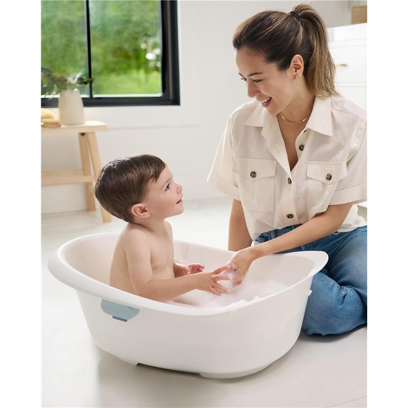 A caregiver interacts with a smiling child in the Skip Hop Wave 4-In-1 Baby Bath Tub filled with bubbles, set in a bright room.