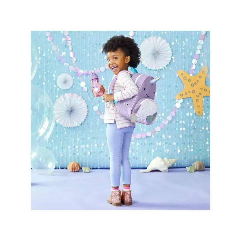 Little girl with curly hair wearing a striped sweater and holding a water bottle, standing with a Skip Hop Narwhal backpack.