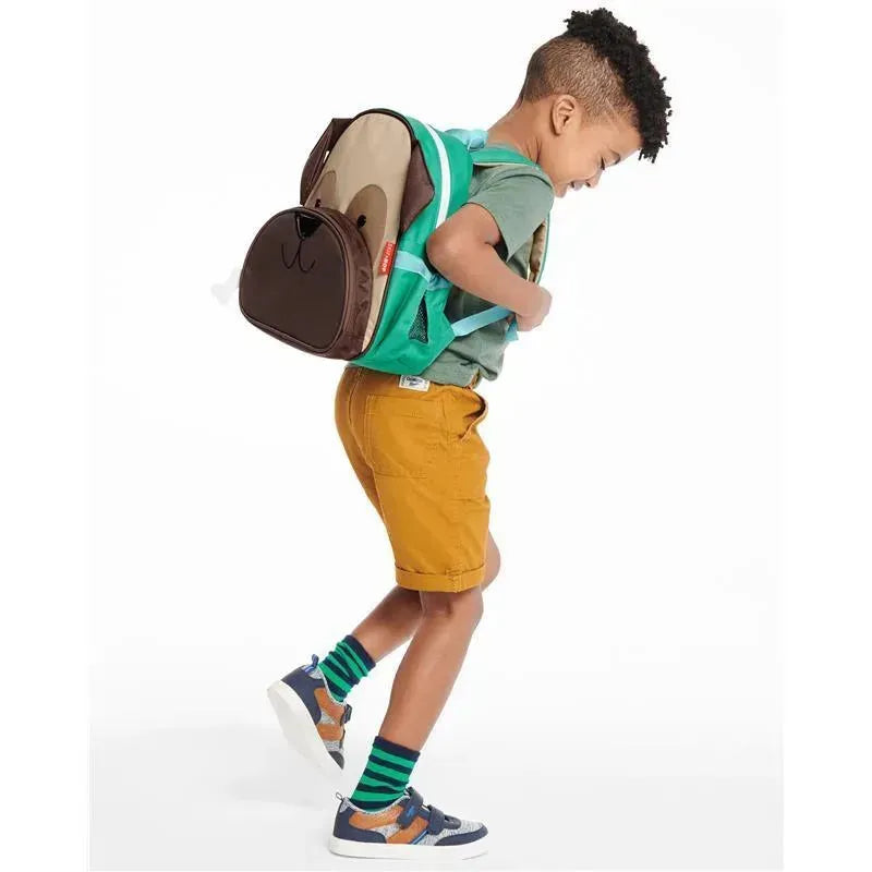 Little boy wearing a green backpack designed as a pug, with yellow shorts and striped socks, smiling while walking.