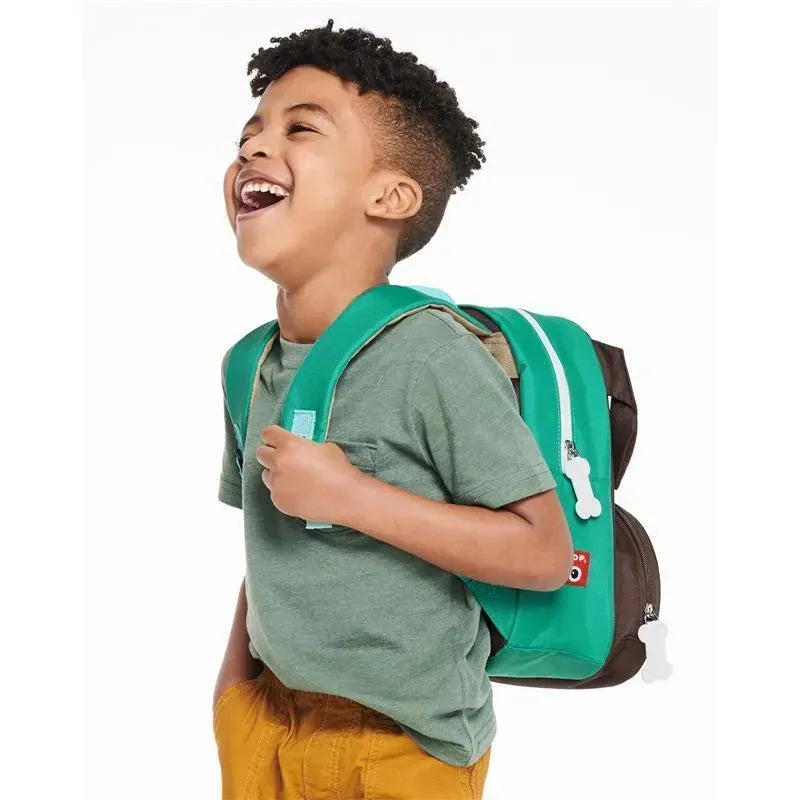 Young boy joyfully wearing a green Skip Hop Zoo Little Kid Backpack featuring a pug design and playful bone-shaped zipper pulls.