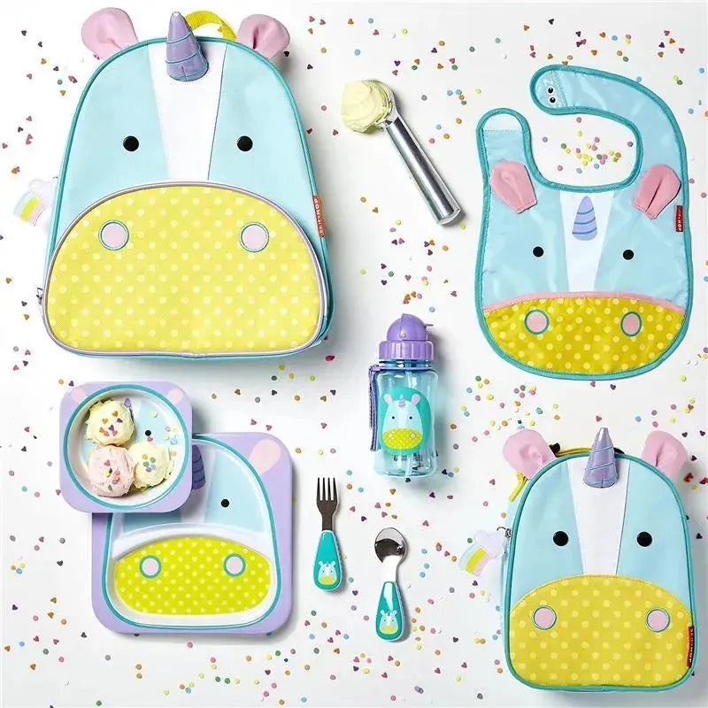 Brightly colored unicorn-themed backpack, bib, plate, and utensils arranged with confetti and ice cream.