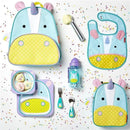 Brightly colored unicorn-themed backpack, bib, plate, and utensils arranged with confetti and ice cream.