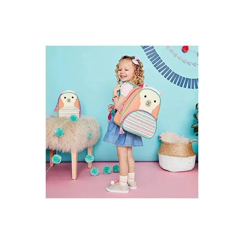 Little girl smiling while wearing a colorful llama-themed backpack, with a similar backpack displayed on a stool nearby.