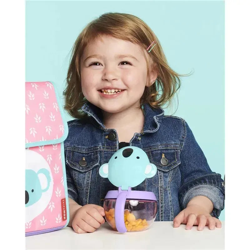 A smiling young girl holds a Skip Hop Zoo Snack Cup shaped like a koala, filled with snacks, next to a matching pink bag.