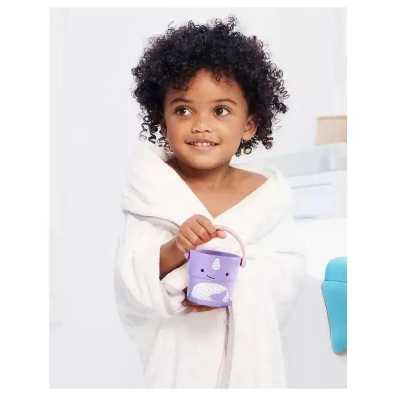 Child in a white bathrobe smiling while holding a purple Skip Hop Zoo Stack & Pour Bucket with a whale design.