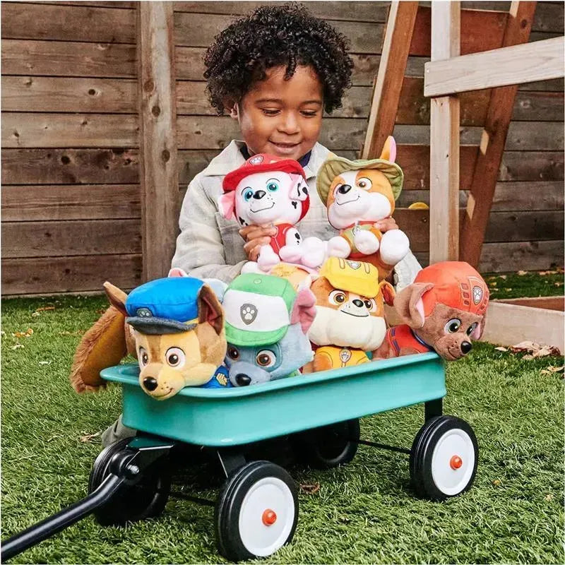 Child smiling while holding plush PAW Patrol characters, seated beside a wagon filled with toys on grass.