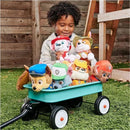 Child smiling while holding plush PAW Patrol characters, seated beside a wagon filled with toys on grass.