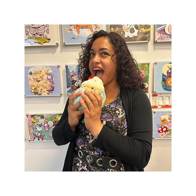 A woman holds a plush cupcake toy, smiling and playfully pretending to eat it, with colorful artwork in the background.