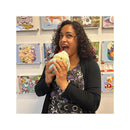 A woman holds a plush cupcake toy, smiling and playfully pretending to eat it, with colorful artwork in the background.